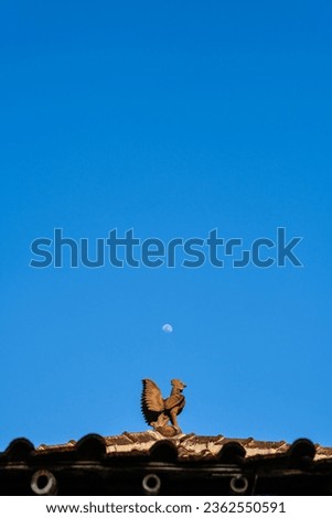 Roof tile decoration in the shape of a chicken with a background of blue sky and moon in the afternoon