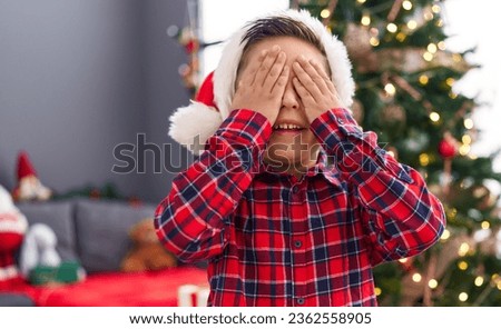 Adorable hispanic boy standing by christmas tree with hands on eyes at home