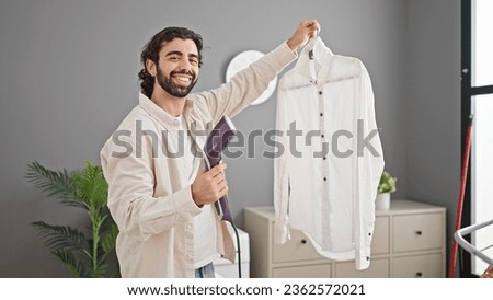 Young hispanic man ironing shirt with vertical iron machine smiling at laundry room
