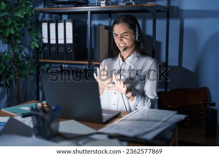 Young brunette woman wearing call center agent headset working late at night disgusted expression, displeased and fearful doing disgust face because aversion reaction. with hands raised  Royalty-Free Stock Photo #2362577869