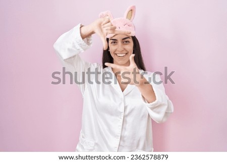 Young brunette woman wearing sleep mask and pajama smiling making frame with hands and fingers with happy face. creativity and photography concept. 