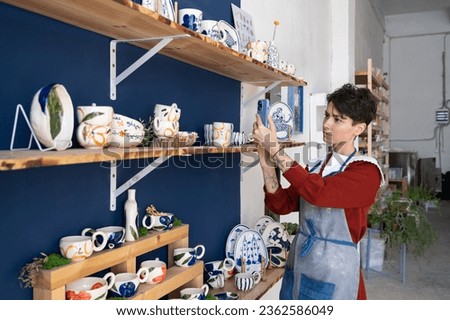 Charismatic Caucasian woman seller in souvenir shop takes pictures of goods at phone for posting on social networks. Successful girl store employee with ceramic tableware shoots video on smartphone