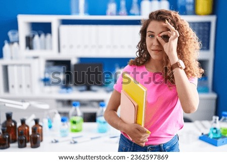 Young caucasian student woman at scientist laboratory smiling happy doing ok sign with hand on eye looking through fingers 