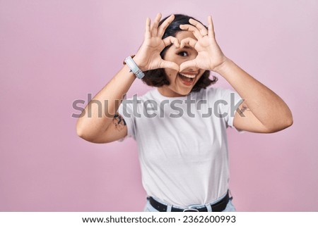 Young hispanic woman wearing casual white t shirt over pink background doing heart shape with hand and fingers smiling looking through sign 