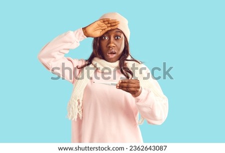 Young woman with cold or flu has fever and takes her temperature. Portrait of sick African American girl wearing warm winter clothes holding thermometer and looking at camera with shocked expression