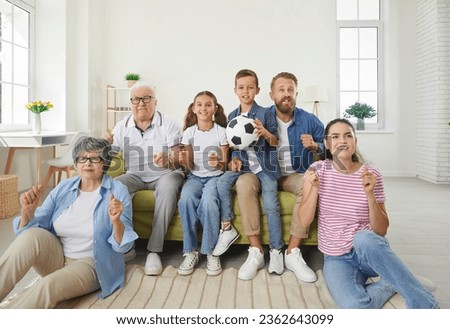 Happy big family watching soccer match on TV in living room at home. Cheerful grandparents, mother, father and two their cute children football fans cheering up support favorite team with soccer ball Royalty-Free Stock Photo #2362643099