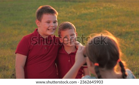Happy little brothers pose to girl taking photo with vintage camera in sunset park. Smiling little boys hug looking in sister camera in field. Brothers pose for family photo to little girl in meadow
