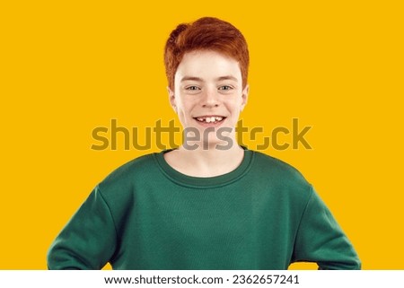 Close-up portrait of smiling, toothless, happy, red-haired, confident teenager dressed in a warm green sweater and looking directly into the camera on an isolated yellow background. Royalty-Free Stock Photo #2362657241