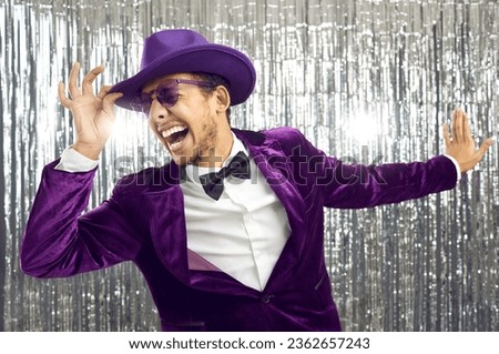 Funny happy excited crazy confident handsome man wearing purple velvet suit, bow tie, glasses and fedora hat having fun, singing loud songs and dancing against shiny foil fringe background at party Royalty-Free Stock Photo #2362657243