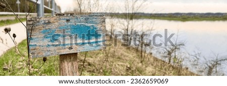 Wooden sign with peeling paint. A sign with old blue paint is embedded in the ground near the river and garden.