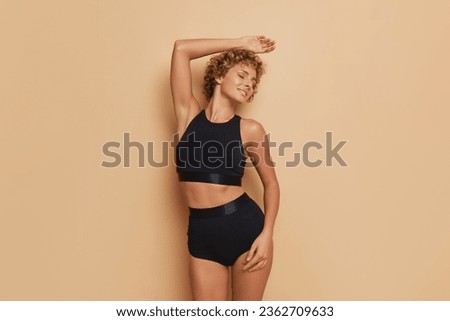 Slim woman in black ergonomic top and pants standing with one hand over her head and closed eyes against beige background, lifestyle concept, copy space