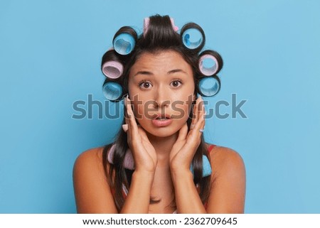 Closeup shot lovely brunette with curlers on her head stands pressing her hands to her face looking surprised, good mood concept, copy space