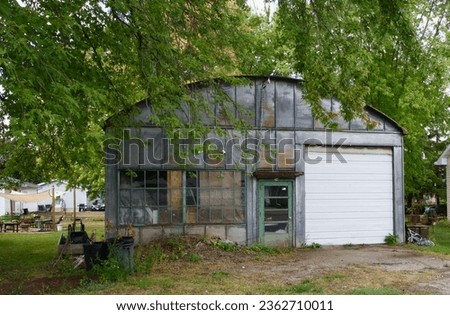 an old abandoned building with a curved roof, large white garage door, green entry door, windows with individual panes on an overgrown lot with a large tree in front Royalty-Free Stock Photo #2362710011