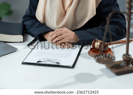Hijab asian muslim female lawyer, lawyer, sitting with hands holding, beautiful smile on legal desk. With laptop, scales and hammer of justice inside the office.