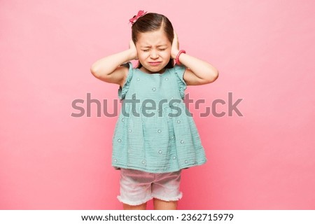 Annoyed elementary little kid feeling upset because of loud noises covering her ears against a pink studio background Royalty-Free Stock Photo #2362715979