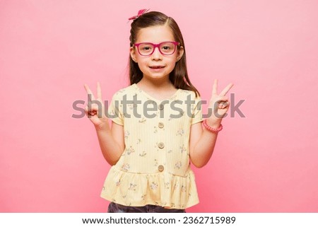 Cheerful little girl making the peace sign and having fun playing while smiling looking happy in a pink studio background Royalty-Free Stock Photo #2362715989