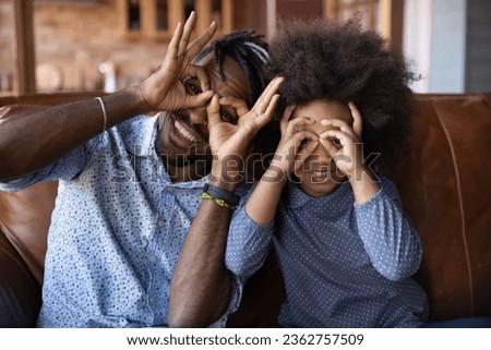 Head shot portrait smiling father with daughter making funny faces, looking at camera through fingers, binoculars glasses eyewear shape gesture, sitting on couch, happy family posing for picture