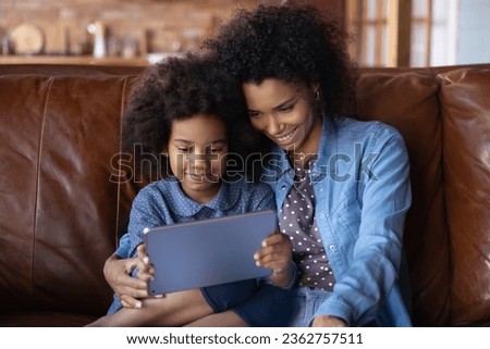 Close up smiling caring African American mother with little daughter using tablet, sitting relaxing on couch at home, happy young mom and 7s girl child spending leisure time with modern device