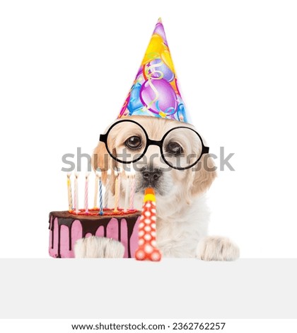 Golden retriever puppy wearing funny eyeglasses and party cap blows into party horn, looks above empty white banner and shows birthday cake with burning candles. isolated on white background