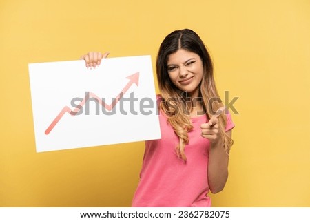 Young Russian girl isolated on yellow background holding a sign with a growing statistics arrow symbol and pointing to the front