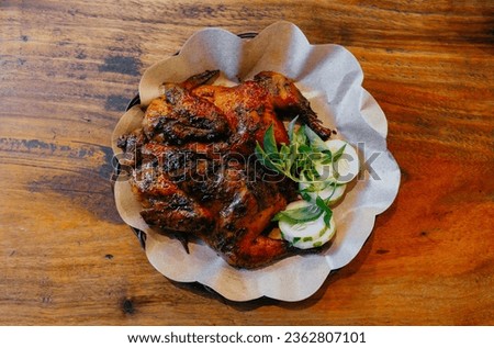 Whole grilled chicken with cucumber slices and basil leaves on a plate with brown greaseproof paper. Horizontal top view with wooden table background. Royalty-Free Stock Photo #2362807101