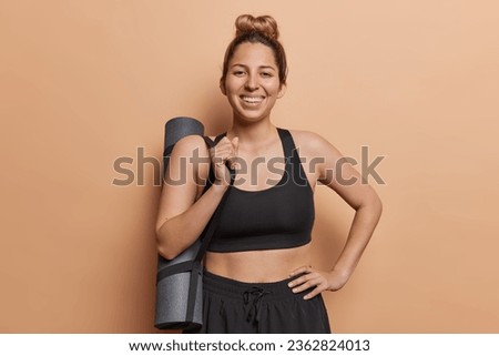 Young beautiful smiling sporty young woman with combed hair dressed in black tracksuit poses with rolled karemar returns home after training in gym has regular workout to keep fit and healthy. Royalty-Free Stock Photo #2362824013