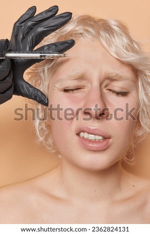 Vertical shot of fair haired woman gets skin lifting injection in forehead frowns face from painful feelings stands shirtless visits beauty clinic isolated over brown background. Anti aging treatment