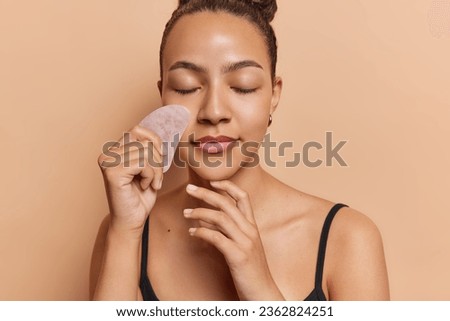 Photo of relaxed dark haired woman massaging face with gua sha stone keeps eyes closed enjoys beauty procedures stands bare shouldered indoor isolated over brown background. Cosmetic natural trend.