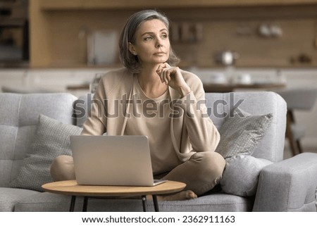 Serious thoughtful mature freelance business woman working at laptop at home, using domestic Internet connection, looking away, thinking, making decision, sitting on sofa at computer, touching chin Royalty-Free Stock Photo #2362911163