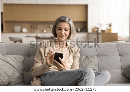 Positive mature grey haired woman enjoying online communication, domestic Internet technology at home, resting on sofa, holding mobile phone, using application for shopping, texting message Royalty-Free Stock Photo #2362911173