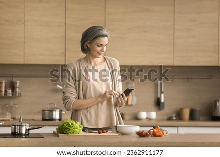 Positive mature chef blogger woman using smartphone over table with organic food ingredients, taking picture, photo, cooking salad, reading online recipe on Internet, smiling