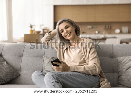 Happy mature naturally grey haired woman holding mobile phone, resting on sofa, looking at camera, posing indoors, smiling, laughing, using digital gadget for domestic Internet communication Royalty-Free Stock Photo #2362911203
