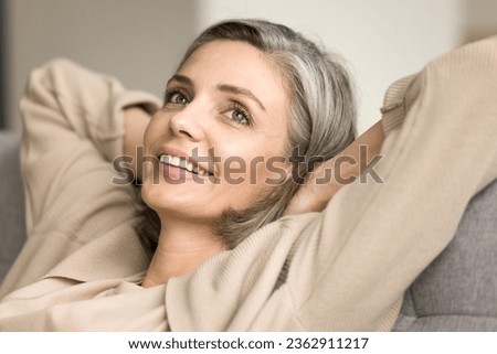 Cheerful beautiful middle aged grey haired woman resting on comfortable sofa, leaning on back with head on hands, looking away with toothy smile, thinking, daydreaming Royalty-Free Stock Photo #2362911217