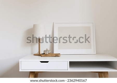 Blank horizontal frame mockup on the white table with the minimalistic table lamp and wooden tray with a silver decorative pumpkin. Modern interior design, home, office desktop.