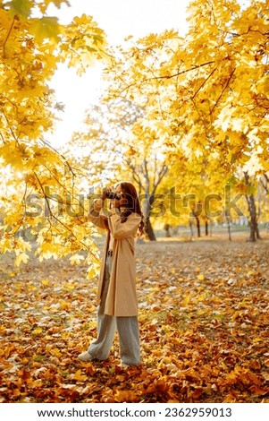 
Smiling woman in a light coat with a retro camera spends time having fun in the autumn forest. Happy mood, leisure time. Lifestyle concept.