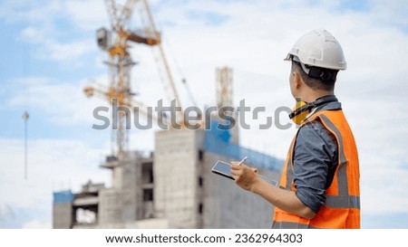 Male site engineer or foreman using digital tablet doing construction site inspection. Asian worker man with orange reflective vest and safety helmet looking at building structure and tower crane
