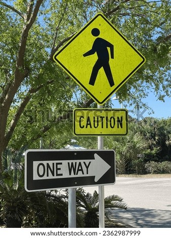 A yellow and black CAUTION sign shows a pedestrian going left and the arrow of a ONE WAY sign going right.   