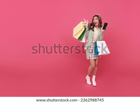 Trendy beautiful young Asian teen woman carrying colorful paper bags shopping online with mobile phone and hand pointing finger isolated on pink banner background with copy space