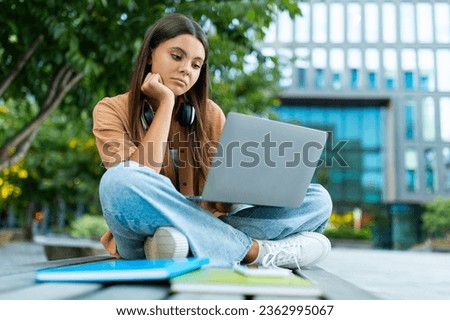Upset young woman university student sitting alone on bench at park, holding laptop computer, doing homework, have difficulties with project or homework. Sad lady feeling lonely, empty space Royalty-Free Stock Photo #2362995067
