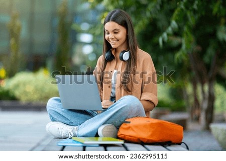 Online education, students lifestyle concept. Cheerful attractive young woman student doing homework at park, sitting on bench, using laptop computer, wireless headphones, copy space Royalty-Free Stock Photo #2362995115