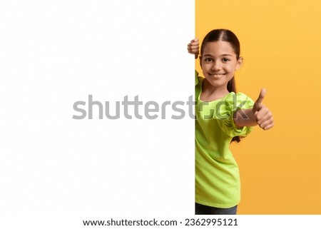 Cute pretty preteen middle eastern girl posing by white blank advertising placard, showing thumb up, nice offer for kids, isolated on yellow background. Blank space mockup for text, advertisement