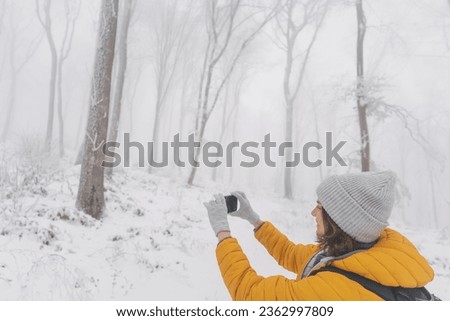 Young caucasian woman in a yellow jacket and hat walking in a snowy forest taking pictures of nature on the phone