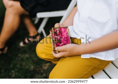 A child, a boy, holds in his hands a glass box with flowers, roses, golden rings with words of love forever at the wedding ceremony. Photo close-up, portrait.