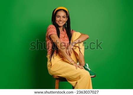 Beautiful young woman with dreadlocs looking at camera while sitting on the chair against green background Royalty-Free Stock Photo #2363037027