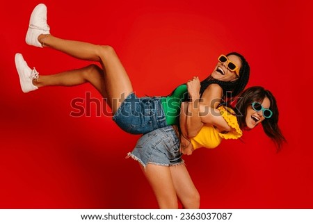 Two playful young women piggybacking each other while having fun against red background together Royalty-Free Stock Photo #2363037087