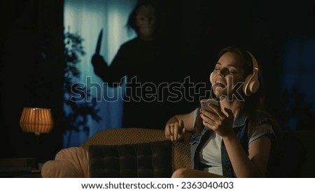 Girl in headphones sitting on the sofa in the living room, listening to music, man maniac in white mask standing with knife near window behind.