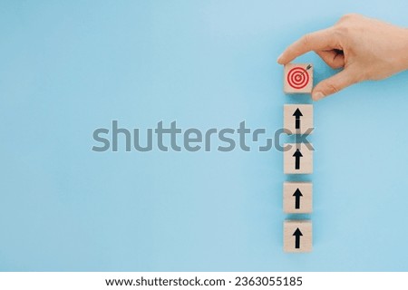 Femail hand holding wooden block with target aiming dart board and arrow up on blue background. Business achievement, objective goal and project progress background, growth value, financial concept