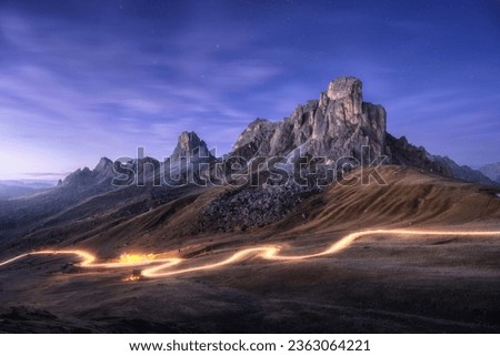 Car light trails on mountain road and high rocks at night in autumn in Passo Giau, Dolomites, Italy. Landscape with blurred light trails, hills, mountain peaks, purple sky with stars in fall at sunset