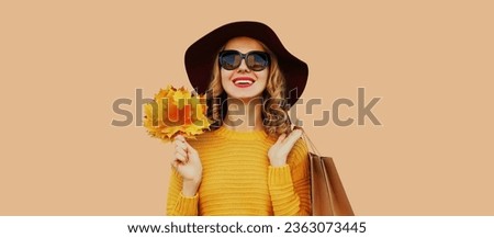 Autumn color style outfit, portrait of beautiful young woman with shopping bag holds yellow maple leaves wearing hat, knitted sweater on beige studio background
