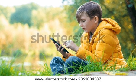 a boy in a yellow jacket sits on the grass in the park, plays a game on his mobile. A 10-year-old child chats outdoors, looks at social networks outside. Royalty-Free Stock Photo #2363084587